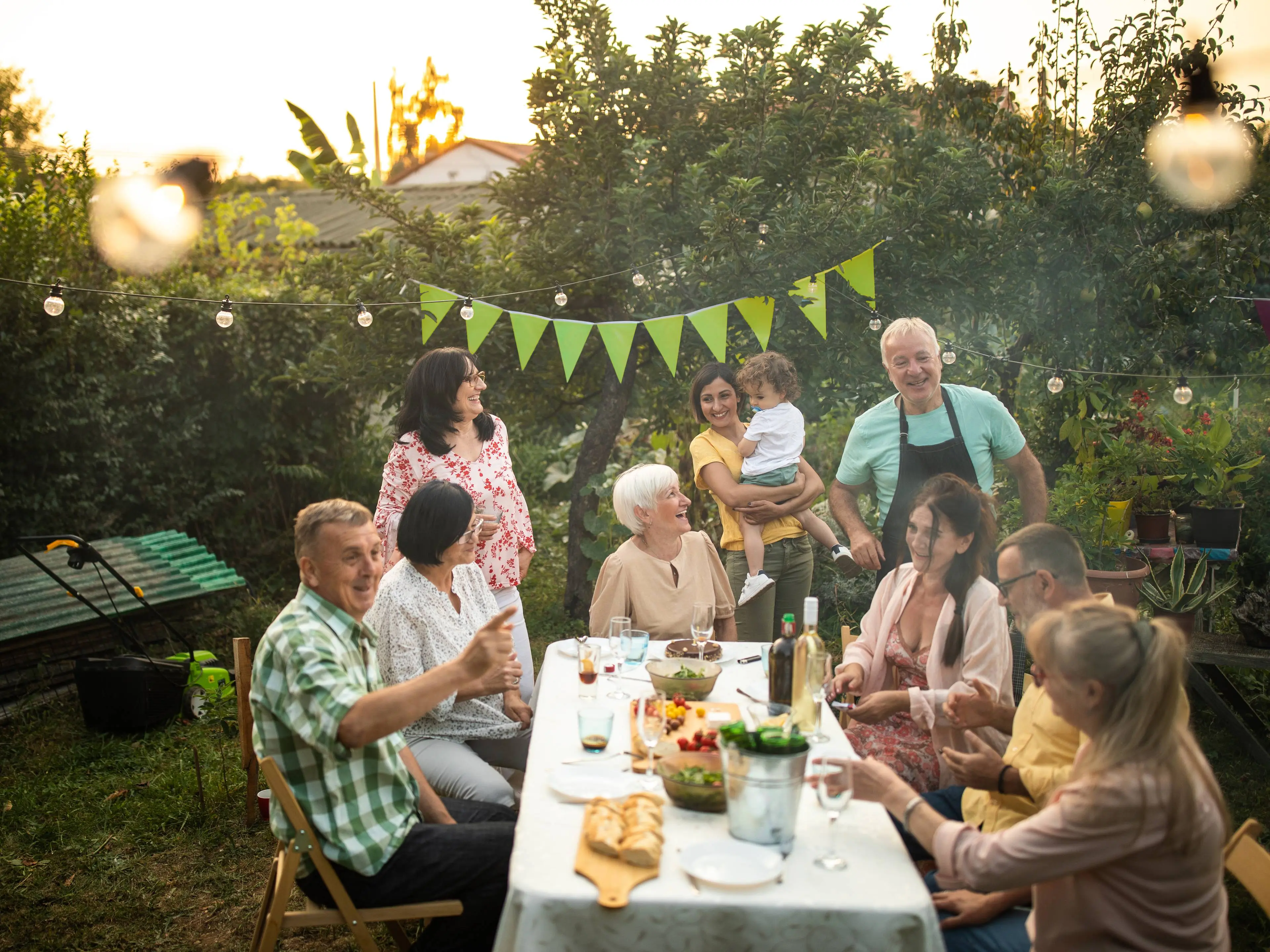 Nachbarn verschiedenen Alters sitzen oder stehen um einen langen Tisch im Garten, lachen und unterhalten sich