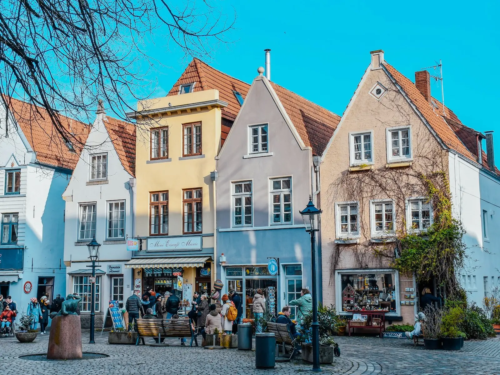 Eine Gruppe von Menschen geht in Bremen auf einer Kopfsteinpflasterstraße spazieren, eine weitere Gruppe steht bei einer Eisdiele an, einige sitzen auf Parkbänken