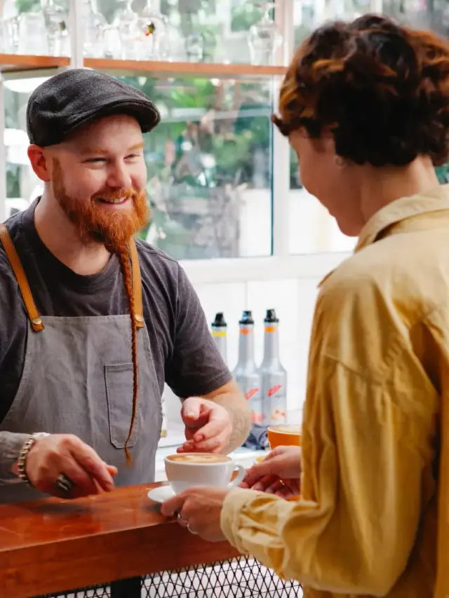 Ein Barista überreicht einer Frau einen Cappuccino am Tresen und lächelt