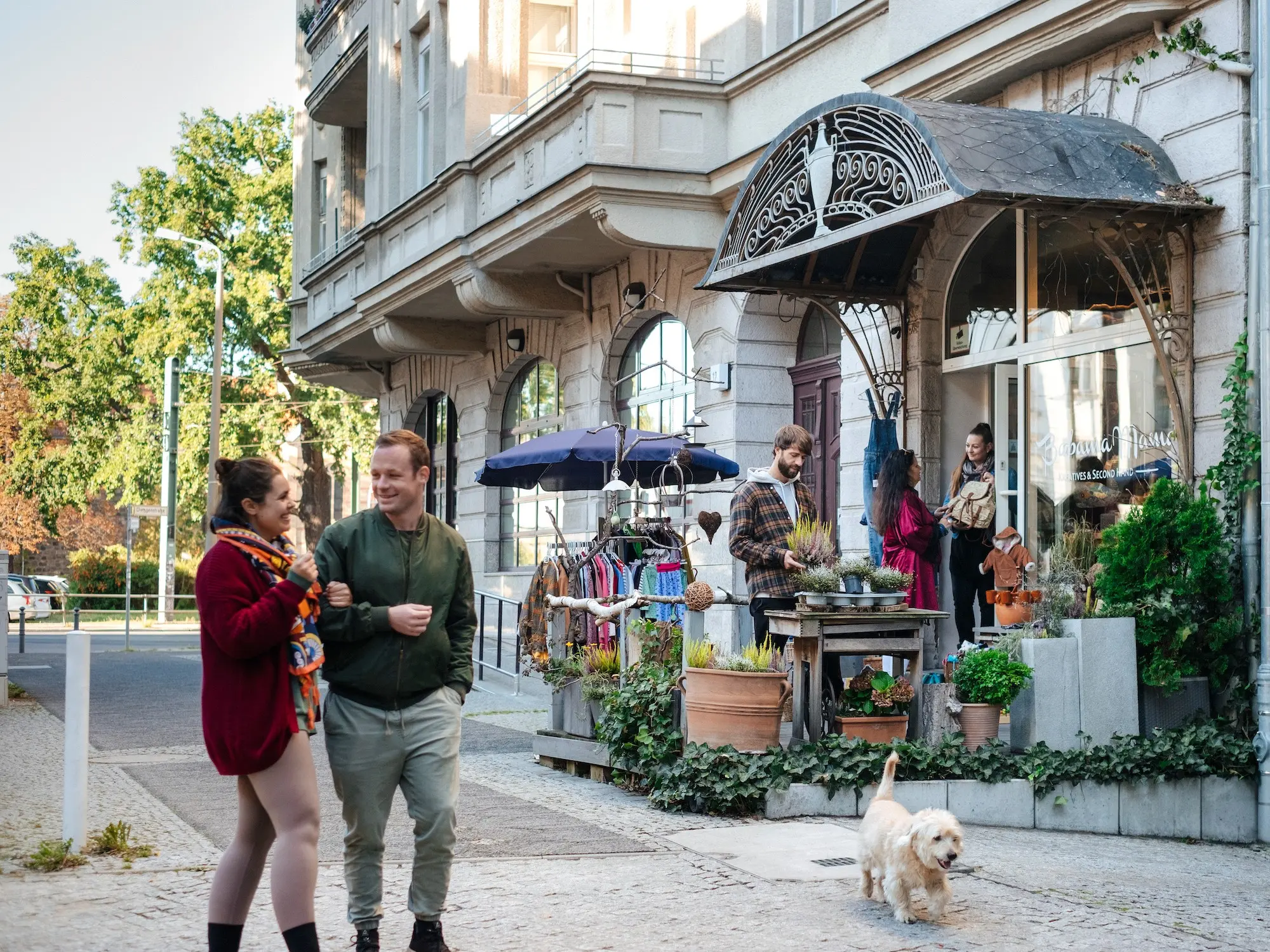 Mehrere Personen schauen sich Artikel außen vor einem Laden in Berlin an, eine Nachbarin und ein Nachbar laufen mit ihrem Hund vorbei